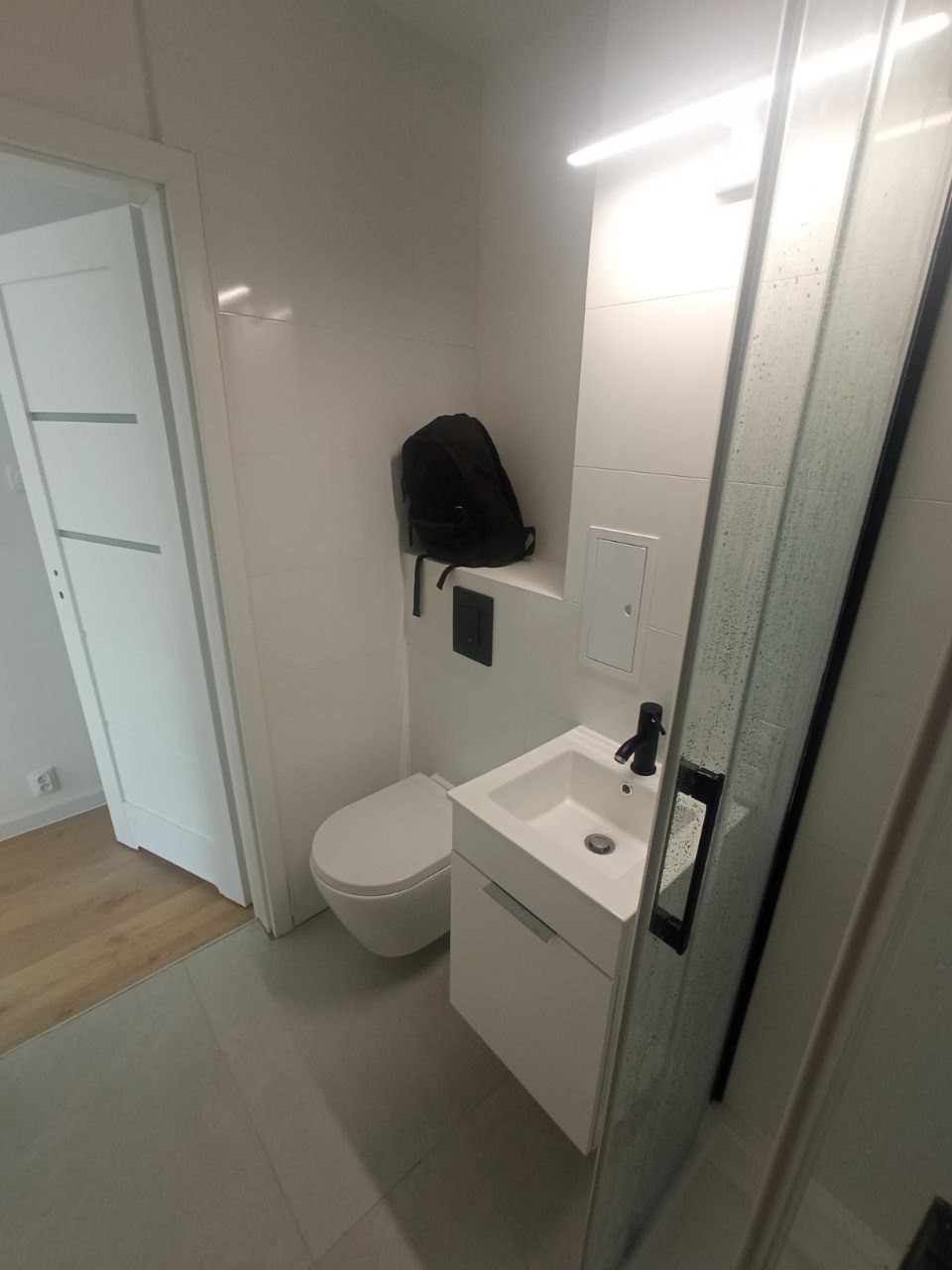 Nowoczesna, minimalistyczna łazienka z białymi płytkami, podwieszaną toaletą, szafką z umywalką i czarną armaturą; plecak leżący na półce nad toaletą.