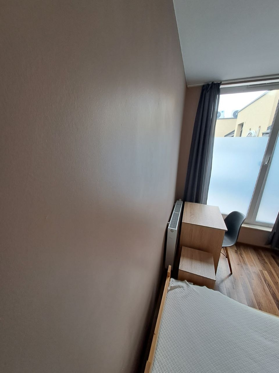 Ściana w pokoju pomalowana na ciepły, beżowy kolor, widoczny grzejnik, biurko z krzesłem przy oknie z zasłoną i częściowo widoczne łóżko.