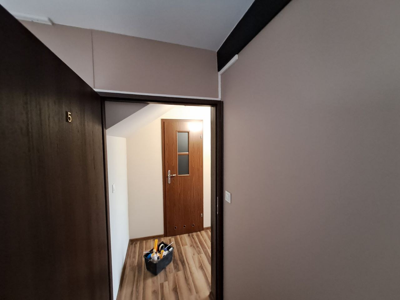 Korytarz po odświeżeniu: świeżo pomalowane ściany w odcieniach beżu, widoczna skrzynka z narzędziami malarskim przy wejściu do mieszkania z brązowymi drzwiami.