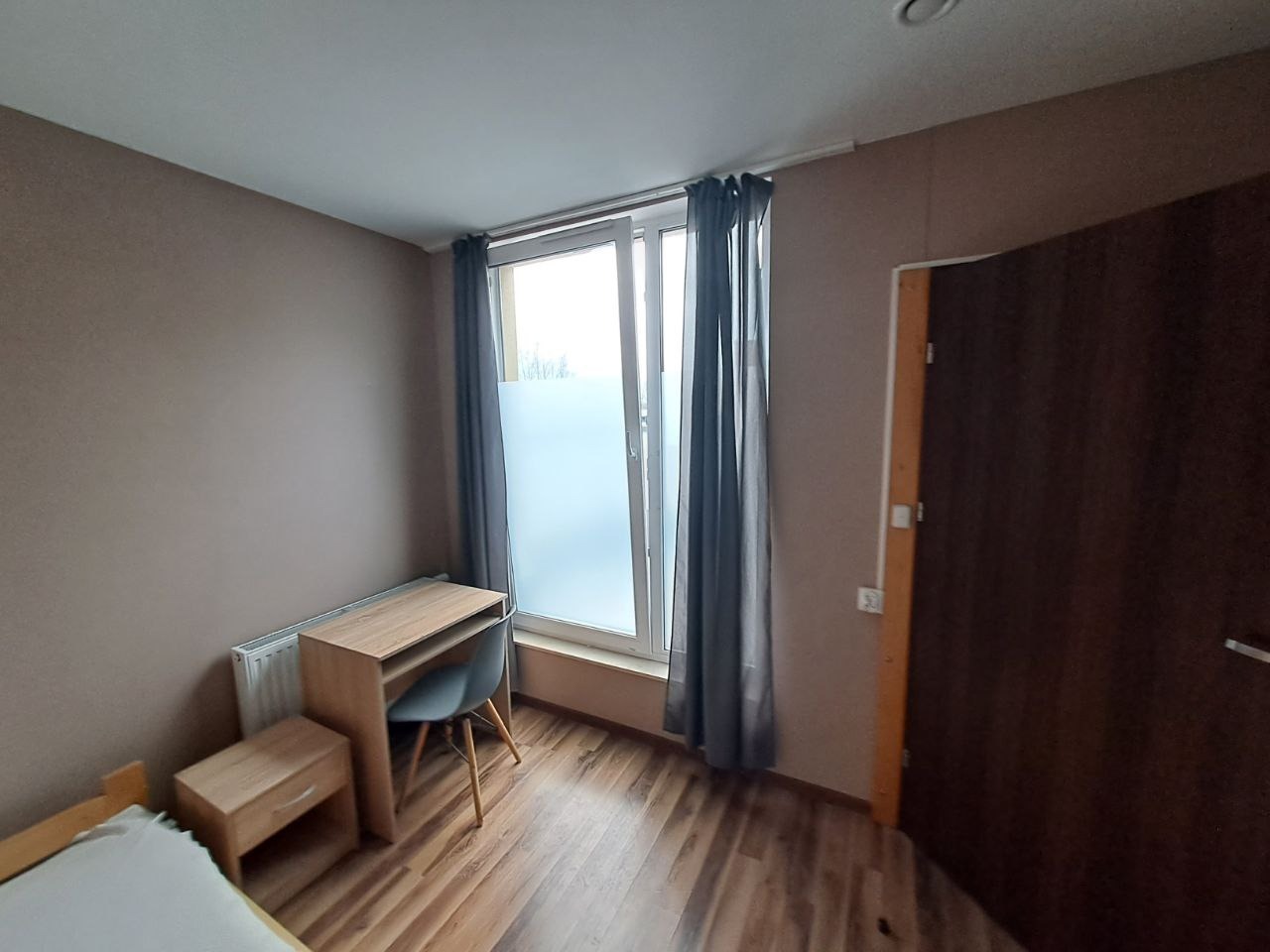 Pokój z drewnianą podłogą, biurkiem, krzesłem i zasłoniętym oknem, ściany w odcieniach brązu, widoczny fragment łóżka.