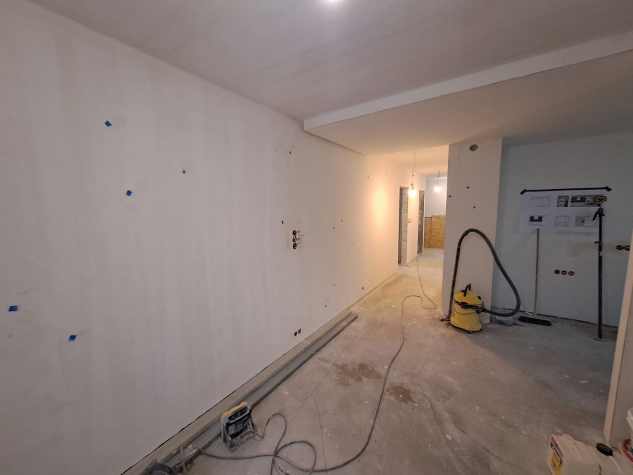 Wnętrze w trakcie prac wykończeniowych: świeżo wyszpachlowane ściany z zaznaczonymi punktami, widoczne przewody elektryczne, rury przy podłodze, odkurzacz przemysłowy i plany architektoniczne...