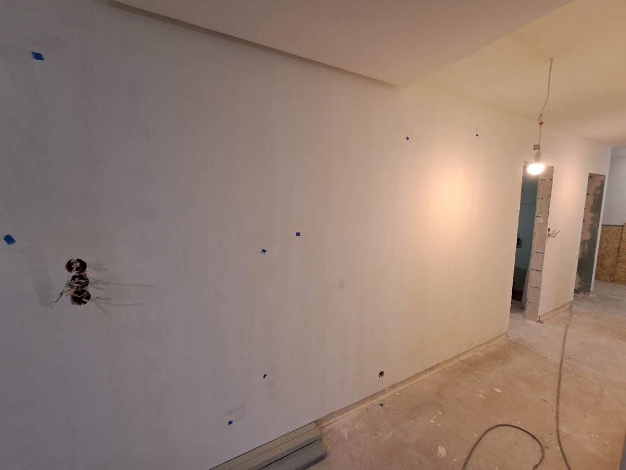 Wnętrze w trakcie prac wykończeniowych: wygładzone ściany z widocznymi otworami na gniazdka elektryczne zabezpieczone taśmą malarską, surowa podłoga, widok na kolejne pomieszczenia przez otwory...