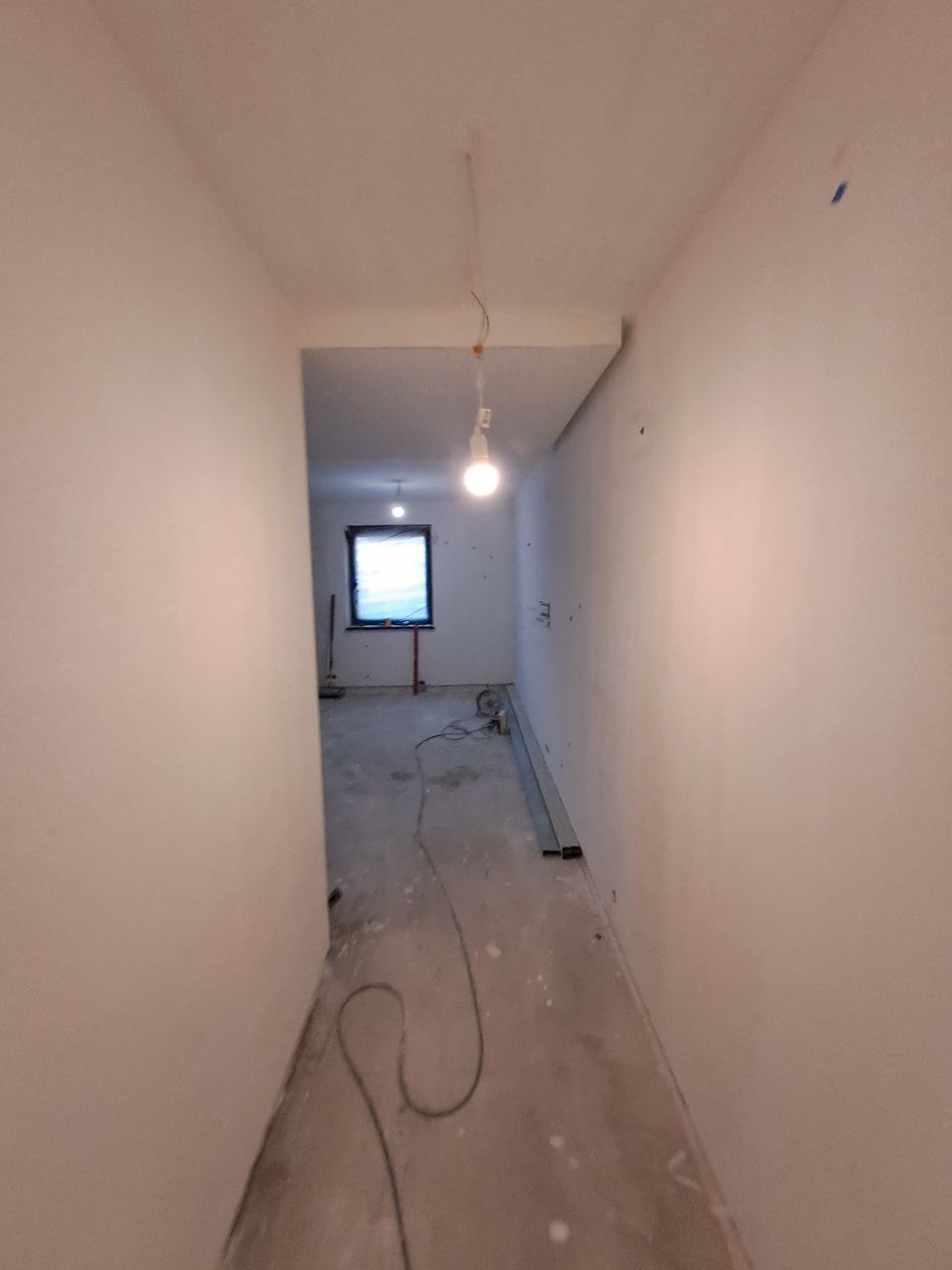 Korytarz w trakcie odświeżania: ściany po szpachlowaniu i malowaniu na biało, surowy beton na podłodze, widoczne kable i tymczasowe oświetlenie żarówką. W głębi pomieszczenia widoczne okno.