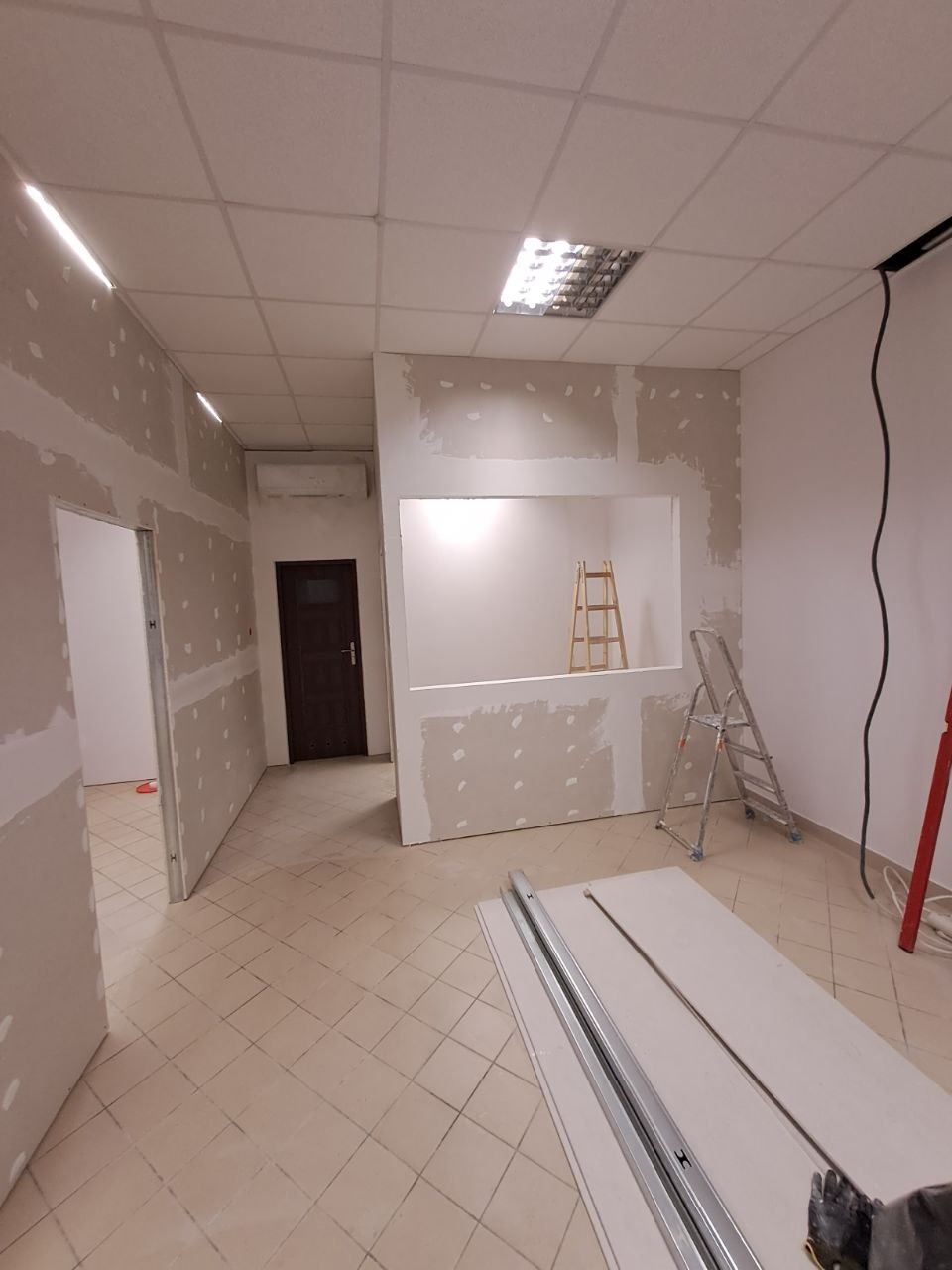 Wnętrze w trakcie montażu płyt kartonowo-gipsowych: widoczne ściany z nałożoną szpachlą, aluminiowe profile na podłodze i drabina w otworze okiennym.