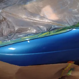 Błyszczący, świeżo polakierowany, niebieski błotnik samochodu, częściowo zakryty folią ochronną i papierem maskującym, zabezpieczony żółtą taśmą malarską.