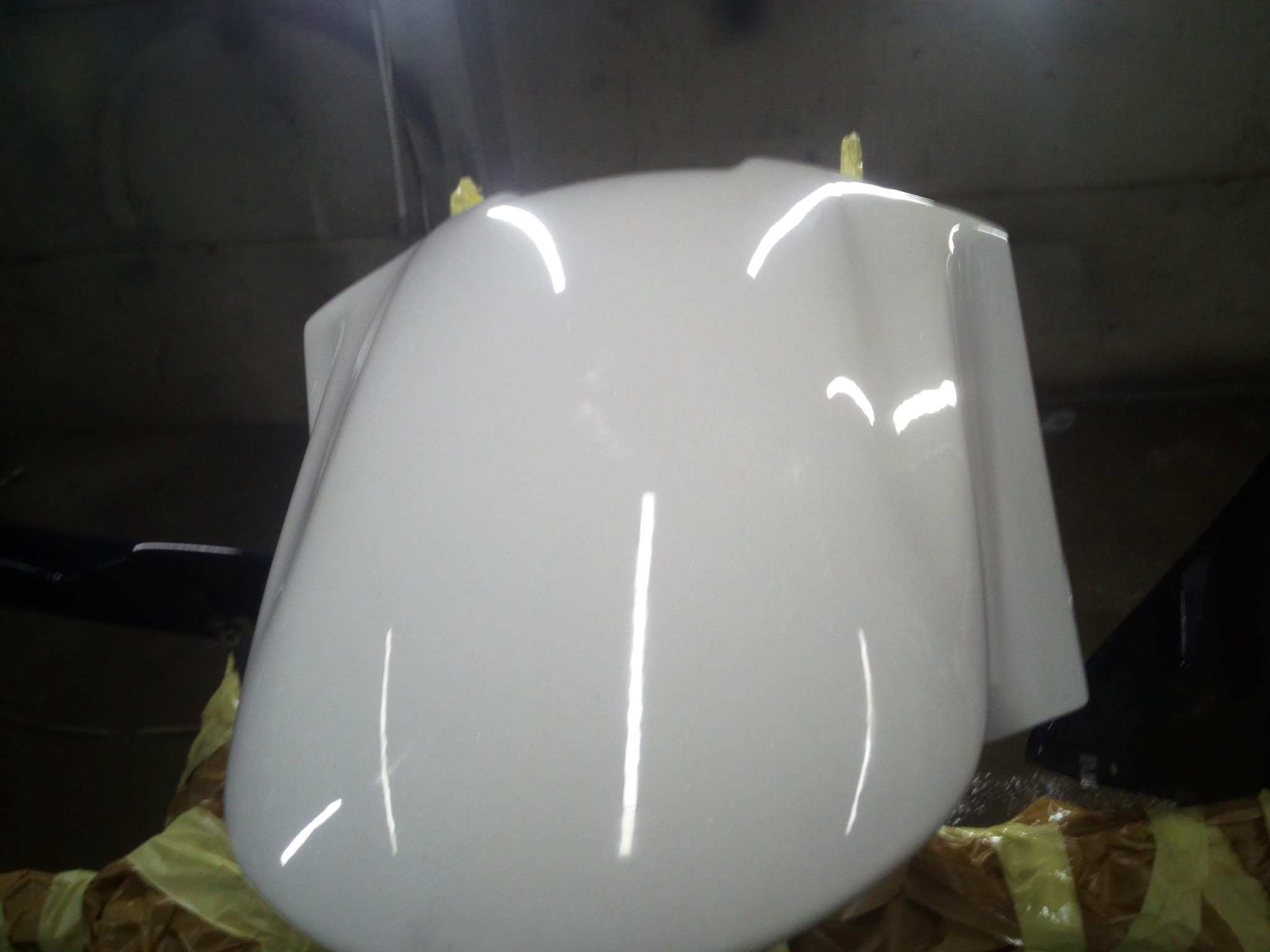 Biała maska samochodu po lakierowaniu, zabezpieczona taśmą malarską, odbijająca światło w ciemnym pomieszczeniu.