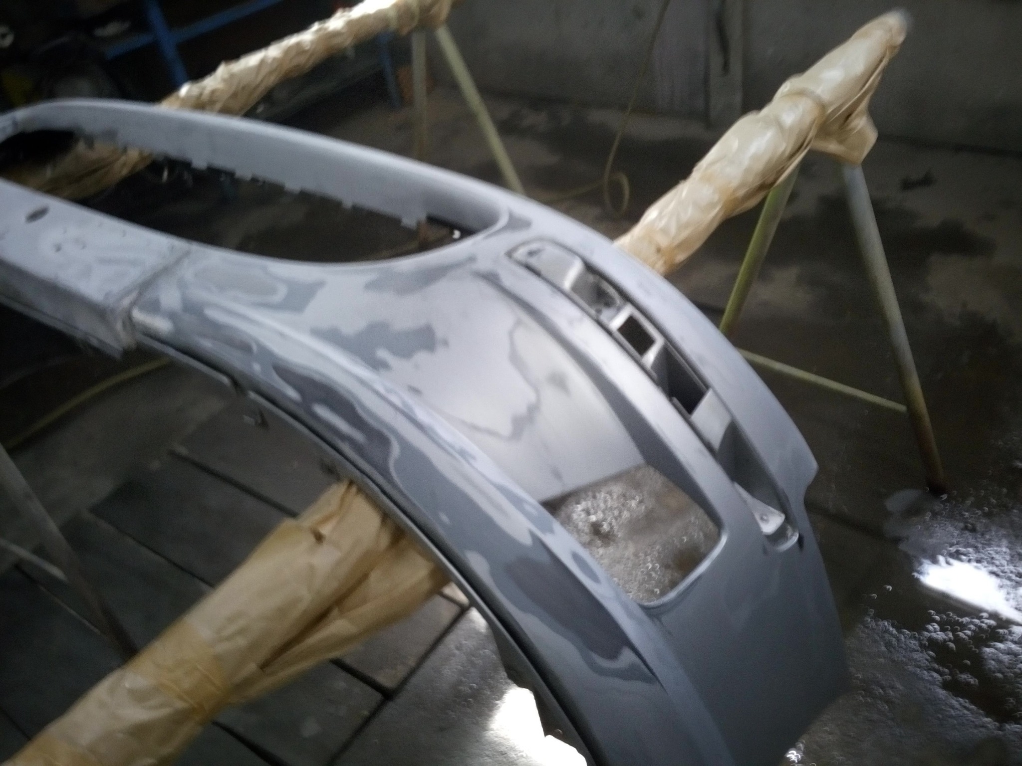 Szary, częściowo wyszlifowany zarysowany zderzak samochodowy na stojaku malarskim w warsztacie, przygotowany do lakierowania. Widoczne ślady szpachli i niedoskonałości powierzchni.