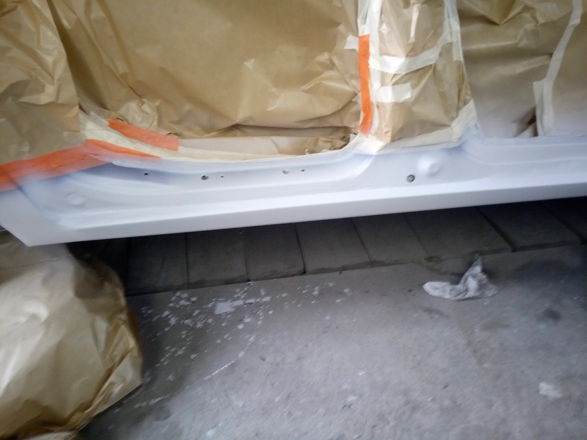 Fragment karoserii samochodu przygotowywany do lakierowania, zabezpieczony papierem i taśmą malarską, widoczne ślady farby na podłodze.