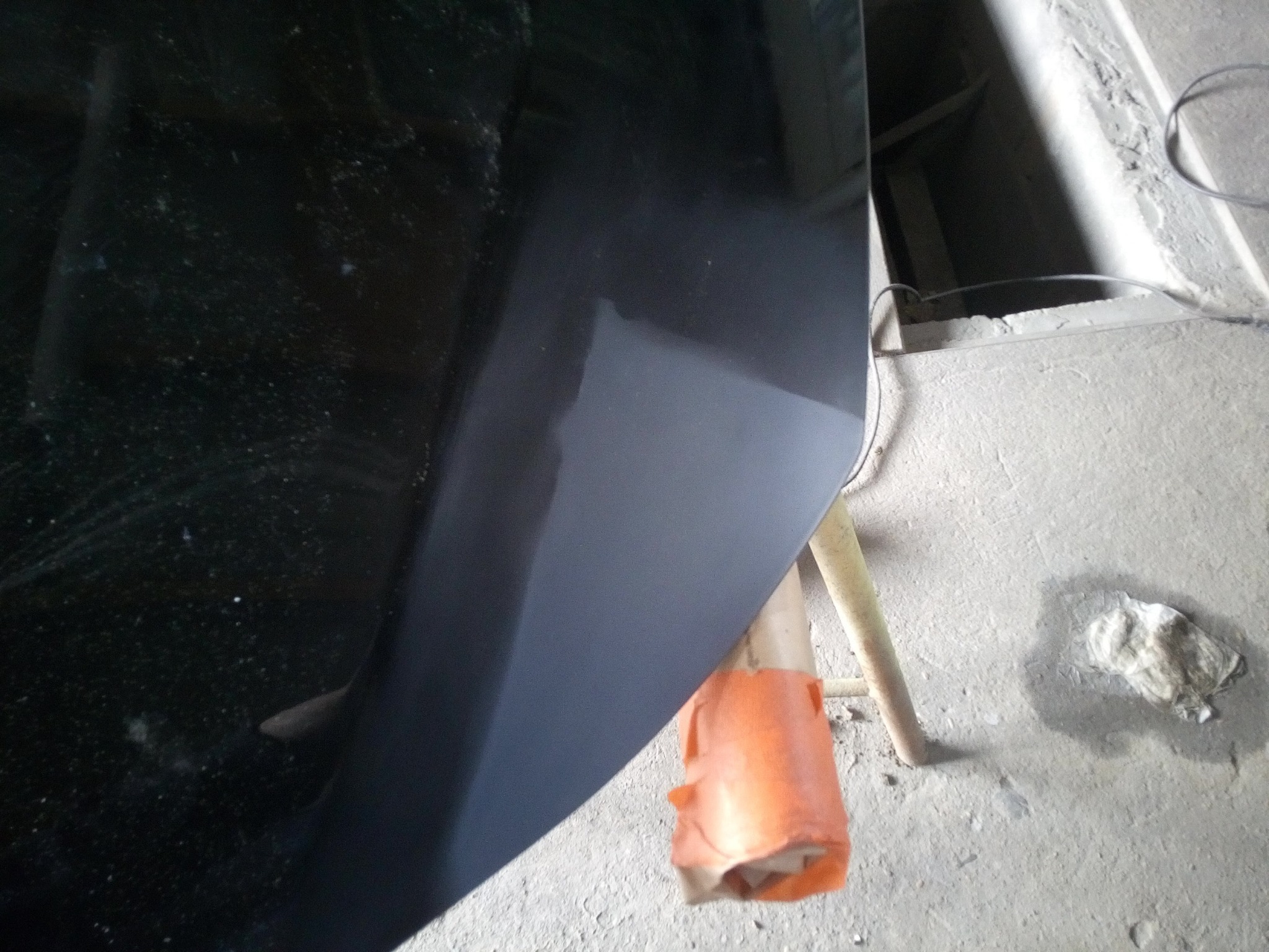 Element karoserii samochodu, prawdopodobnie maska, w trakcie szpachlowania lub przygotowywania do lakierowania. Widoczne niedoskonałości na powierzchni i fragment warsztatowego otoczenia.