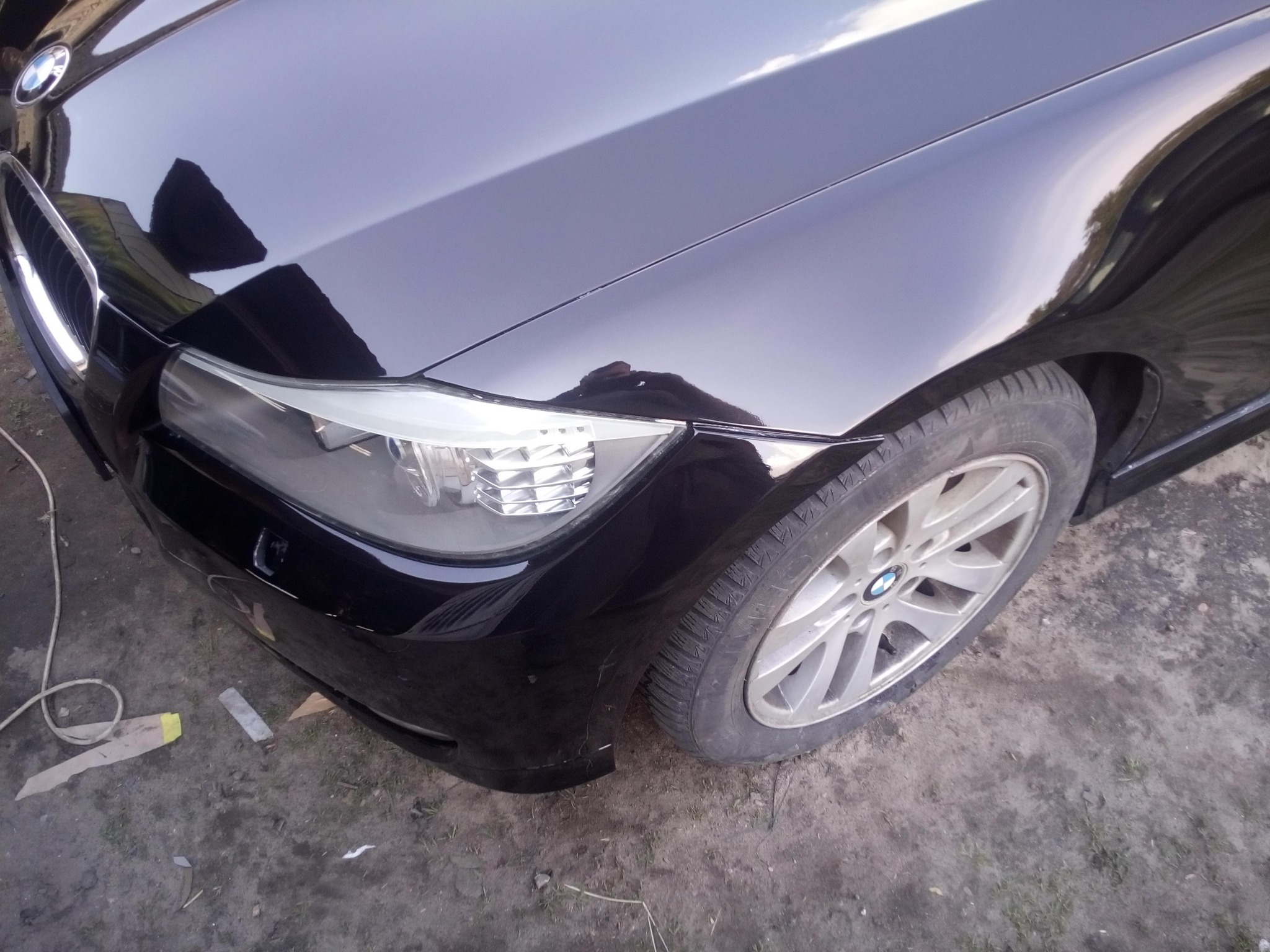 Fragment czarnego samochodu marki BMW; widoczny błotnik, reflektor i opona. Na ziemi leżą kawałki papieru i przewód.