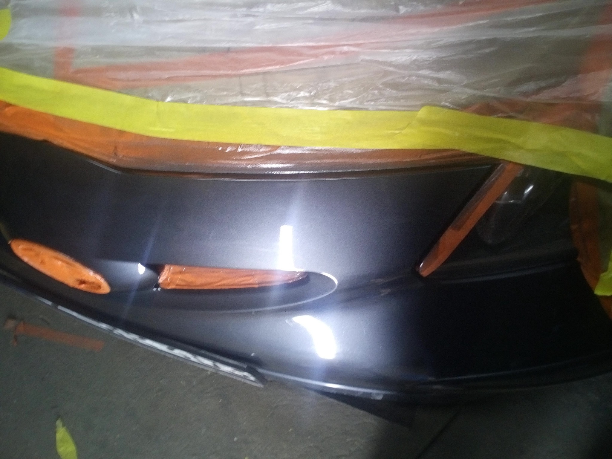 Przygotowany do lakierowania, osłonięty folią i taśmą, fragment błotnika samochodowego w szarym kolorze, widoczne światło odblaskowe na powierzchni.