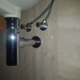 Błyszczący chromowany syfon umywalkowy z podłączonymi elastycznymi wężami i zaworami odcinającymi na beżowej ścianie z naturalnego kamienia.