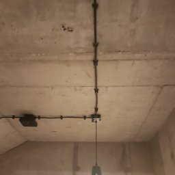HUBERT - Złota Rączka - - Betonowy sufit z natynkową instalacją elektryczną, czarne kable przymocowane klamrami, wisząca lampa w stylu industrialnym z zielonym kloszem.