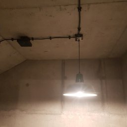 HUBERT - Złota Rączka - - Surowy betonowy sufit z widoczną instalacją elektryczną w czarnych rurkach, puszką połączeniową i zawieszoną lampą z zielonym kloszem, świecącą jasnym światłem.