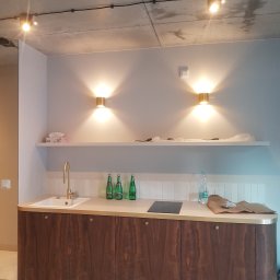 Hubert Kajtaniak - Nowoczesna kuchnia z drewnianymi szafkami, złotą armaturą i oświetleniem punktowym na suficie z betonu architektonicznego, widoczne butelki na blacie i akcesoria montażowe.