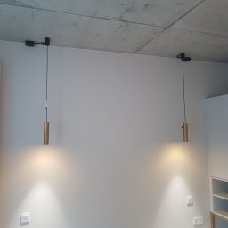 HUBERT - Złota Rączka - - Dwie złote lampy wiszące cylindryczne z czarnymi przewodami zamocowane do surowego betonowego sufitu, widoczne gniazdka elektryczne i włączniki na ścianie.
