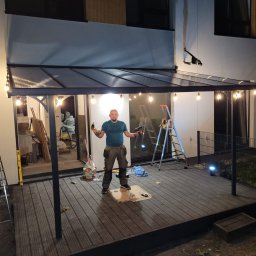 Hubert Kajtaniak - Mężczyzna z wiertarkami w rękach, stojący na kartonie na drewnianym tarasie pod zadaszeniem z oświetleniem, drabiny w tle, prawdopodobnie po montażu konstrukcji.