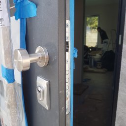 Hubert Kajtaniak - Szare drzwi wejściowe z metalową klamką i zamkiem, zabezpieczone niebieską taśmą malarską, widok na wnętrze w trakcie remontu z sylwetką osoby na tle okna.