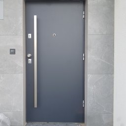 Hubert Kajtaniak - Grafitowe drzwi wejściowe z długim, pionowym uchwytem, zamontowane w szarej, kamiennej elewacji, widoczny fragment kostki brukowej i narożnik kartonu.