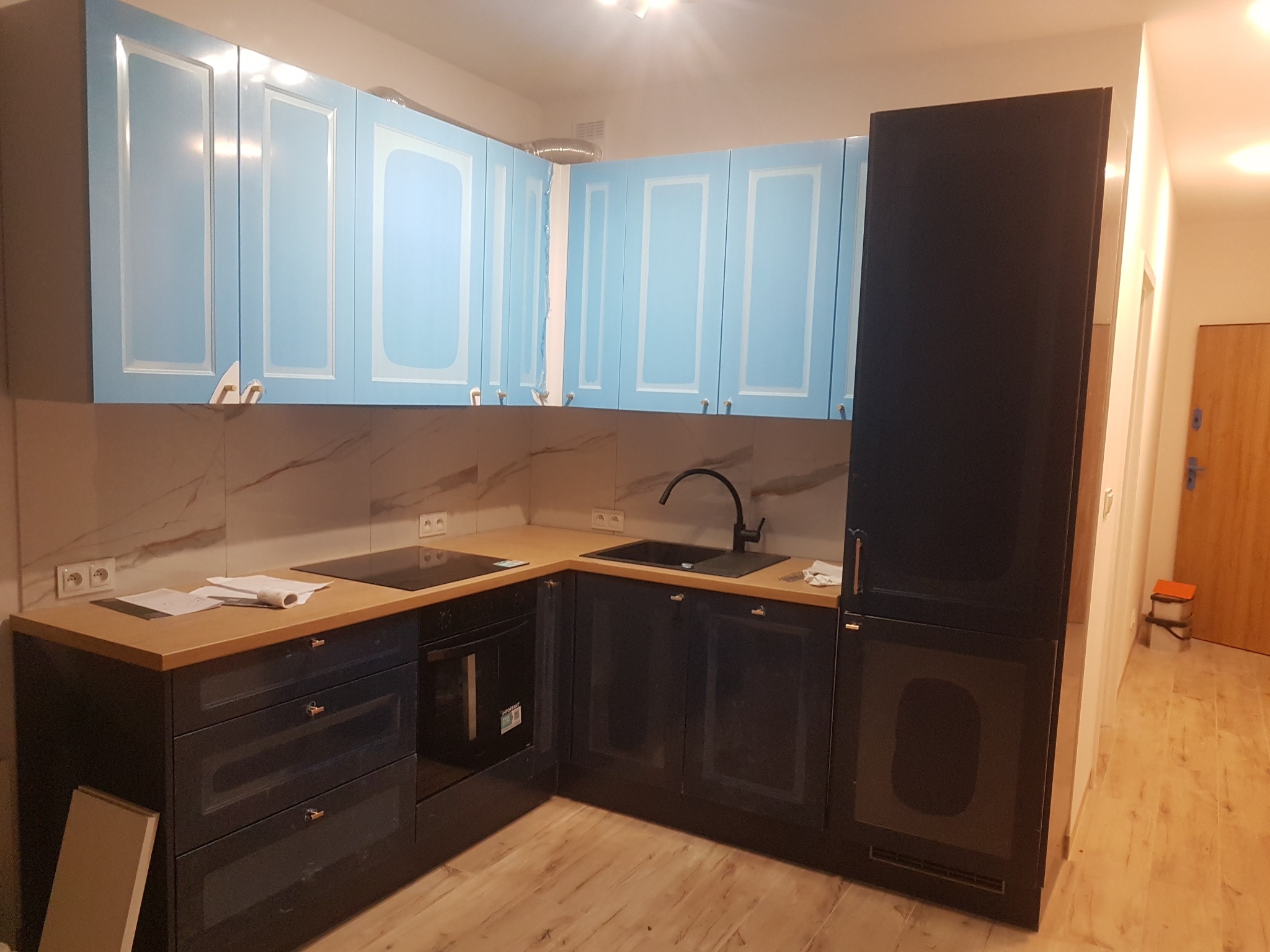 Zmontowana kuchnia z błękitnymi szafkami wiszącymi z frezowanymi frontami, czarnymi szafkami dolnymi i drewnianym blatem, czarnym zlewem i baterią, płytą indukcyjną oraz zabudowaną lodówką.
