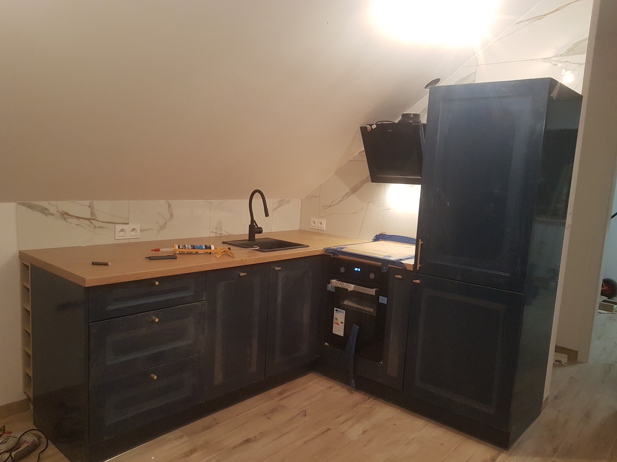 Nowoczesna, granatowa zabudowa kuchenna pod skosem, z drewnianym blatem i czarną armaturą, widok na zamontowany piekarnik i okap, na blacie leżą narzędzia i akcesoria montażowe.