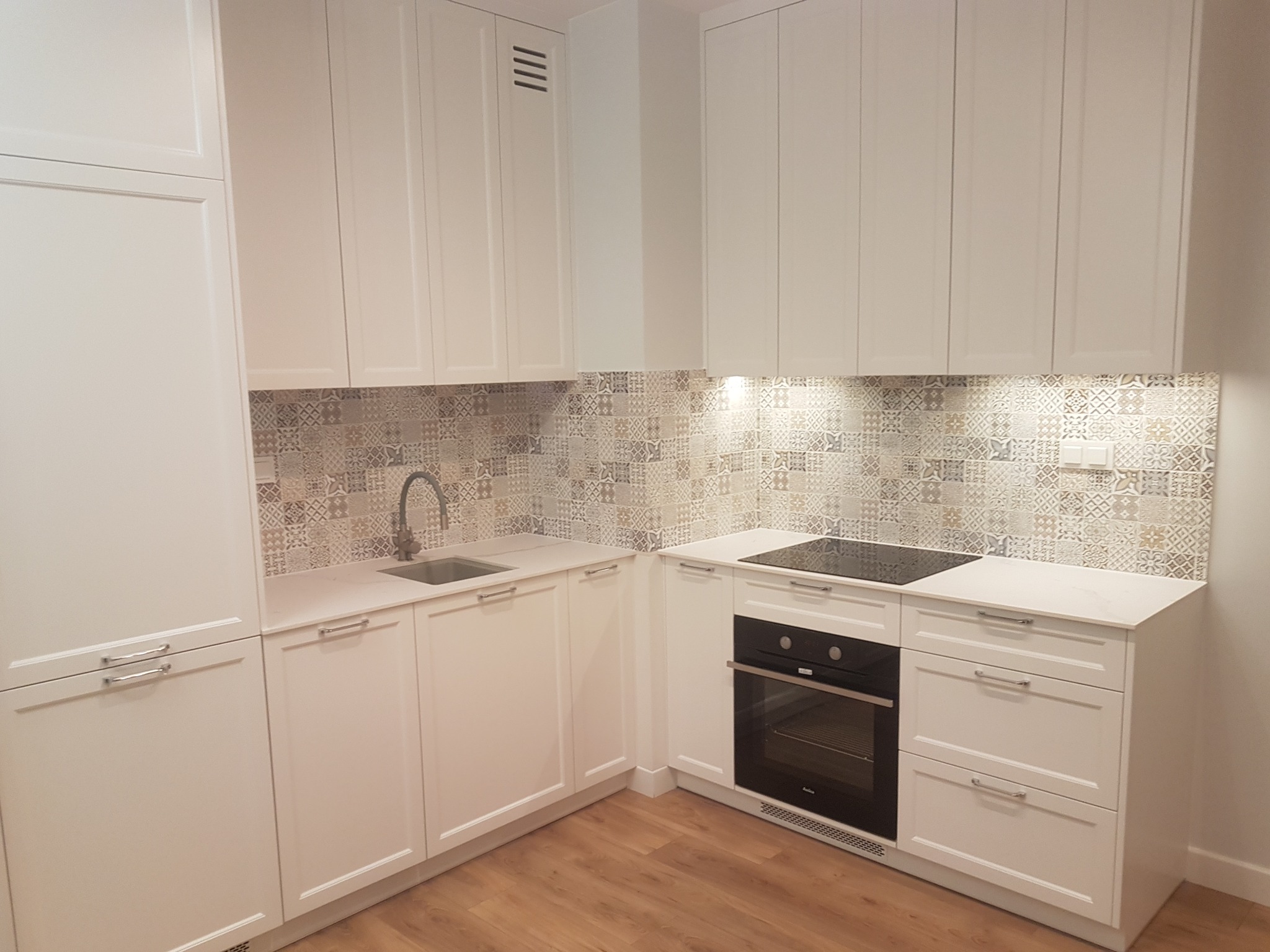 Nowoczesna, jasna kuchnia z białymi szafkami, wzorzystą mozaiką nad blatem, płytą indukcyjną i piekarnikiem w zabudowie, z drewnianą podłogą