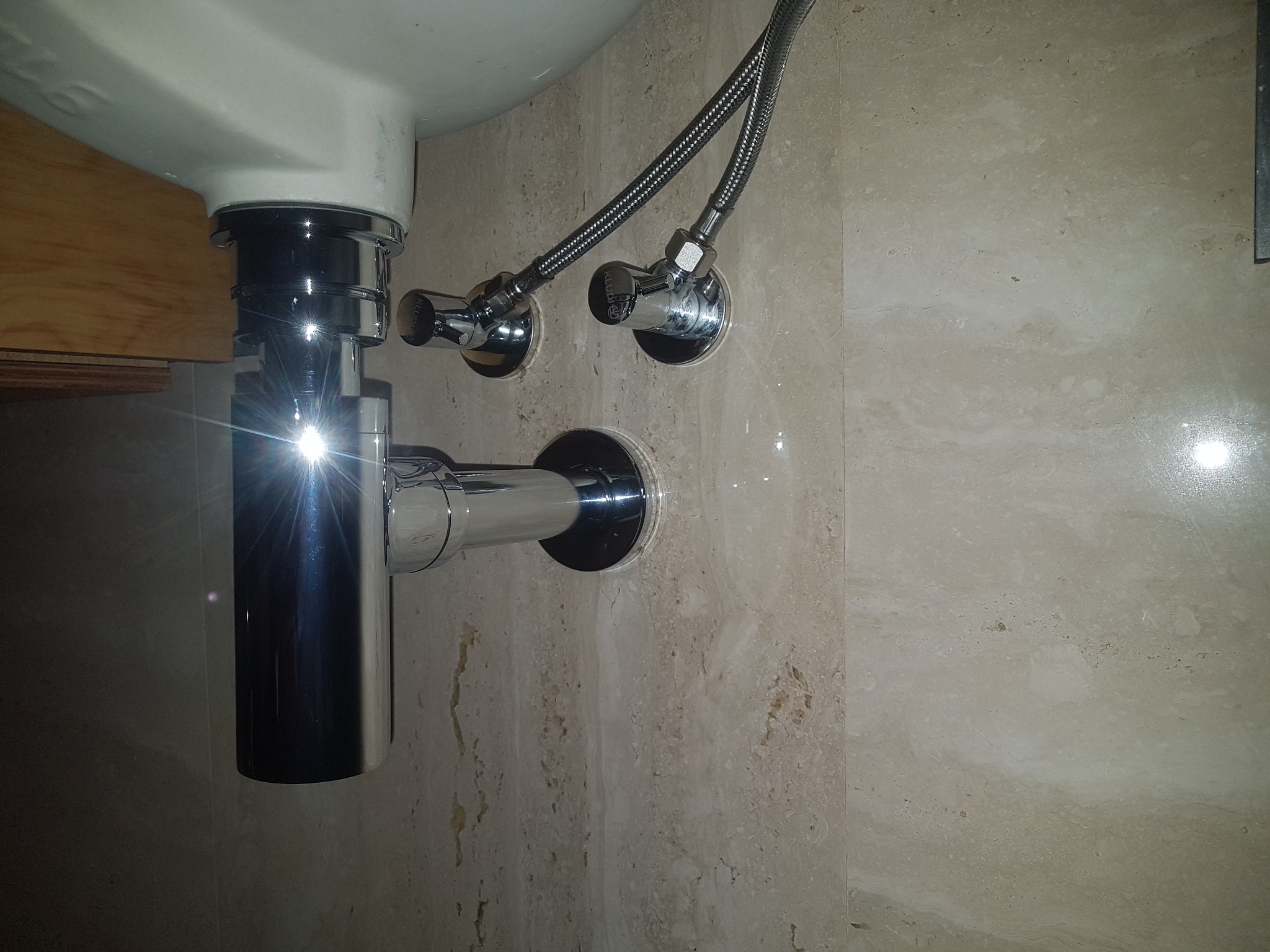 Błyszczący chromowany syfon umywalkowy z podłączonymi elastycznymi wężami i zaworami odcinającymi na beżowej ścianie z naturalnego kamienia.