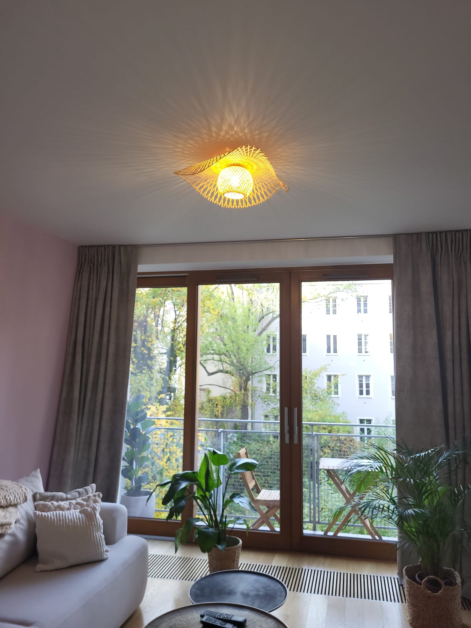 Wnętrze salonu z widokiem na balkon, centralnym punktem jest dekoracyjna lampa wisząca z abażurem z wikliny, rzucająca promieniujące cienie na sufit. W tle widoczne okno balkonowe z drewnianą ramą...