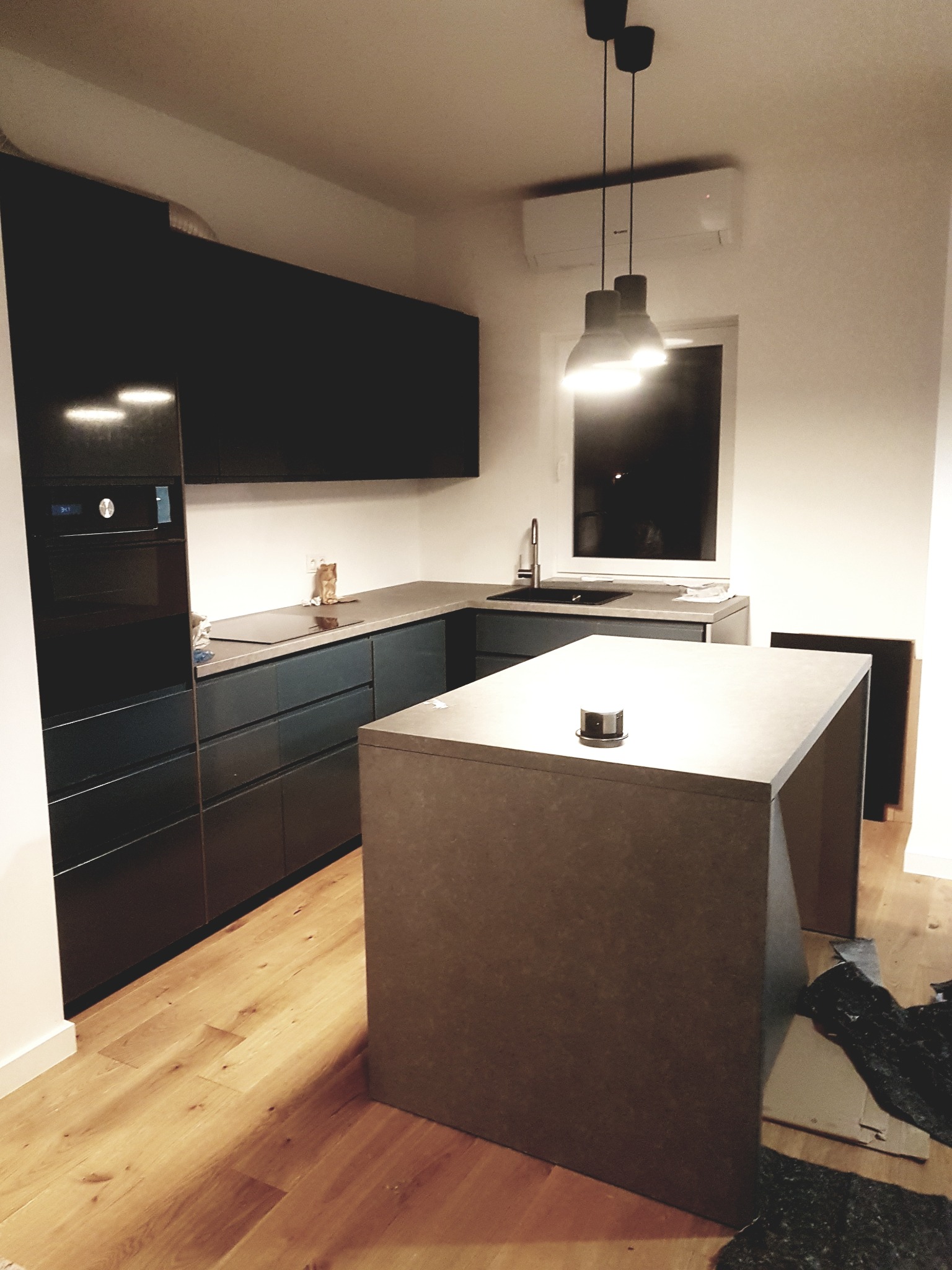 Nowoczesna, ciemna kuchnia z wyspą kuchenną i drewnianą podłogą, oświetlona wiszącymi lampami, z widocznym fragmentem instalacji klimatyzacji pod sufitem.