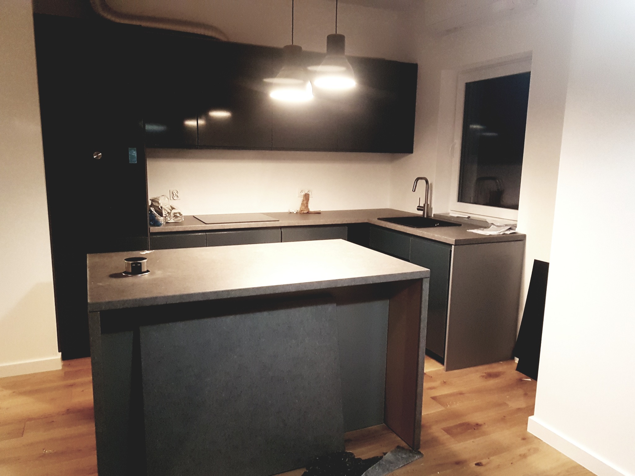 Nowoczesna kuchnia z ciemnymi, matowymi szafkami, wyspą kuchenną z wbudowanym gniazdkiem i drewnianą podłogą, oświetlona dwoma wiszącymi lampami.