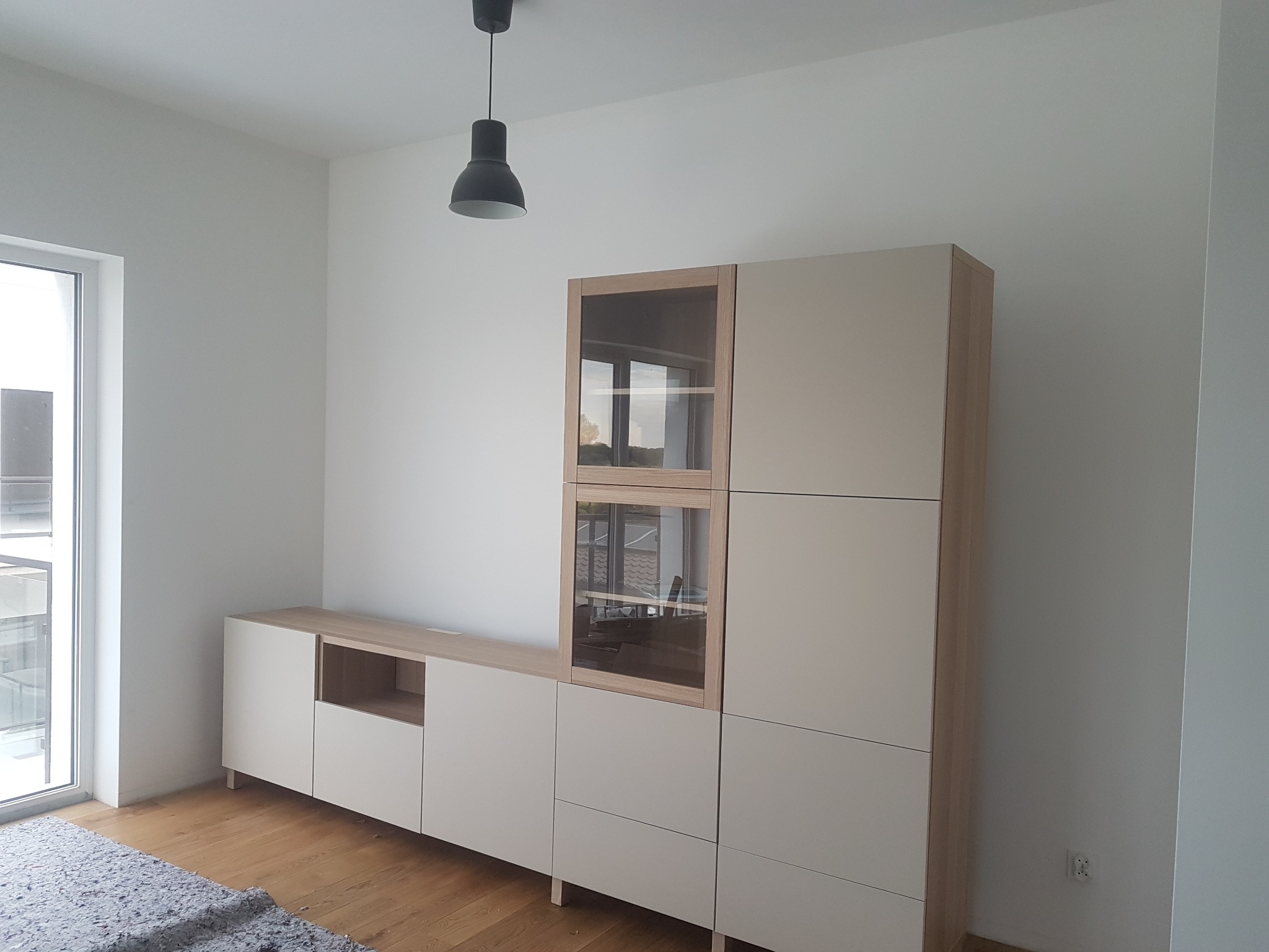 Jasny, nowoczesny zestaw mebli z przeszkloną witryną i szafkami, ustawiony na drewnianej podłodze w minimalistycznym wnętrzu z widoczną lampą sufitową.