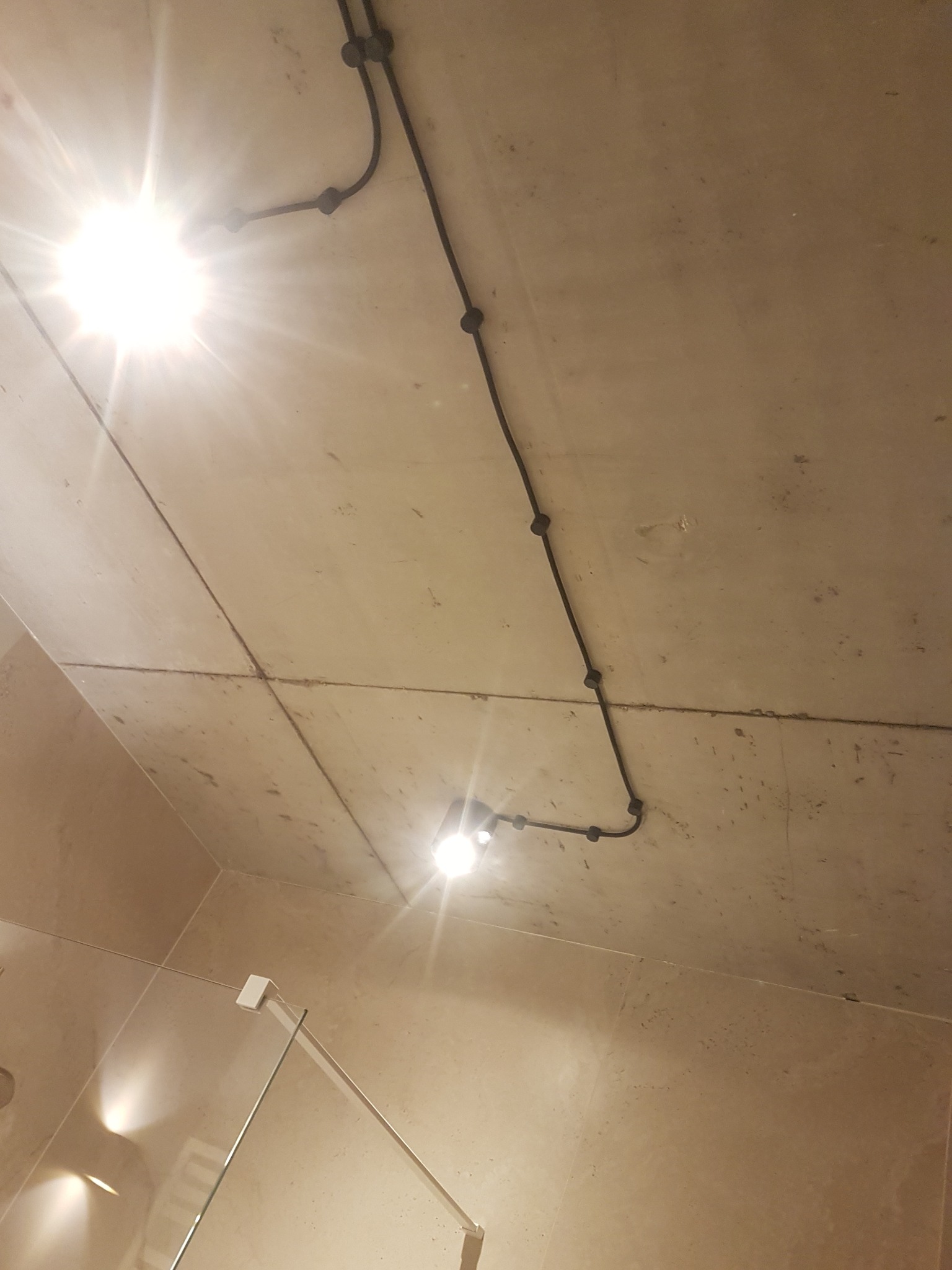 Niefortunny widok: Czarny kabel elektryczny prowadzony po suficie łazienki z widocznymi punktami mocowania i dwoma żarówkami, jasne światło.