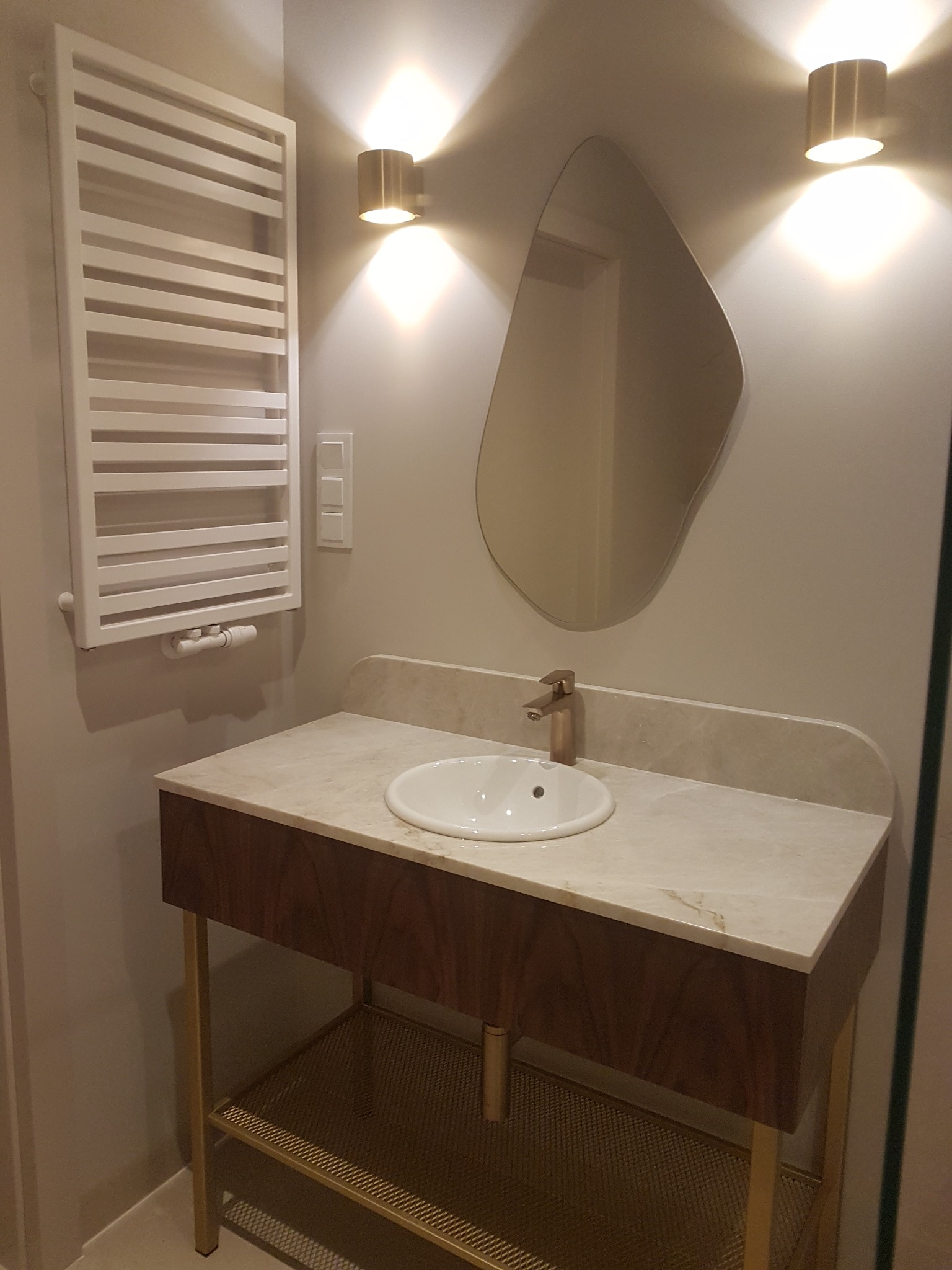 Nowoczesna łazienka z umywalką nablatową, marmurowym blatem, szafką z ciemnego drewna na złotych nóżkach, designerskim lustrem o nieregularnym kształcie i grzejnikiem drabinkowym.