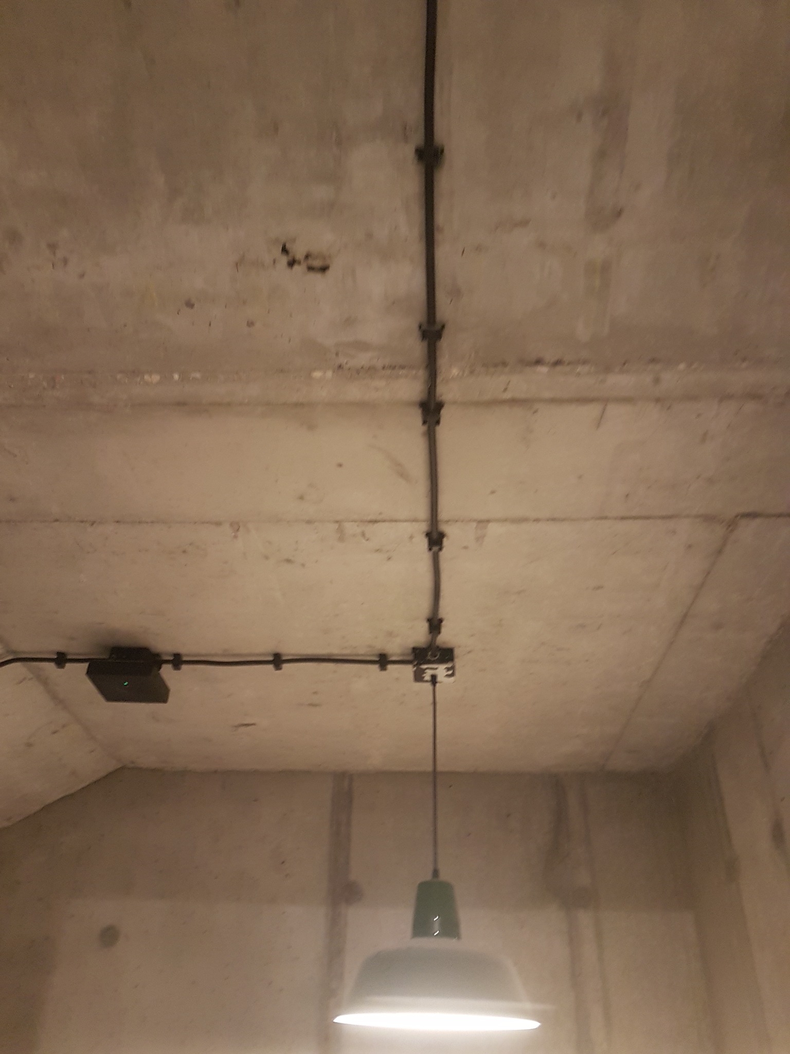 Betonowy sufit z natynkową instalacją elektryczną, czarne kable przymocowane klamrami, wisząca lampa w stylu industrialnym z zielonym kloszem.