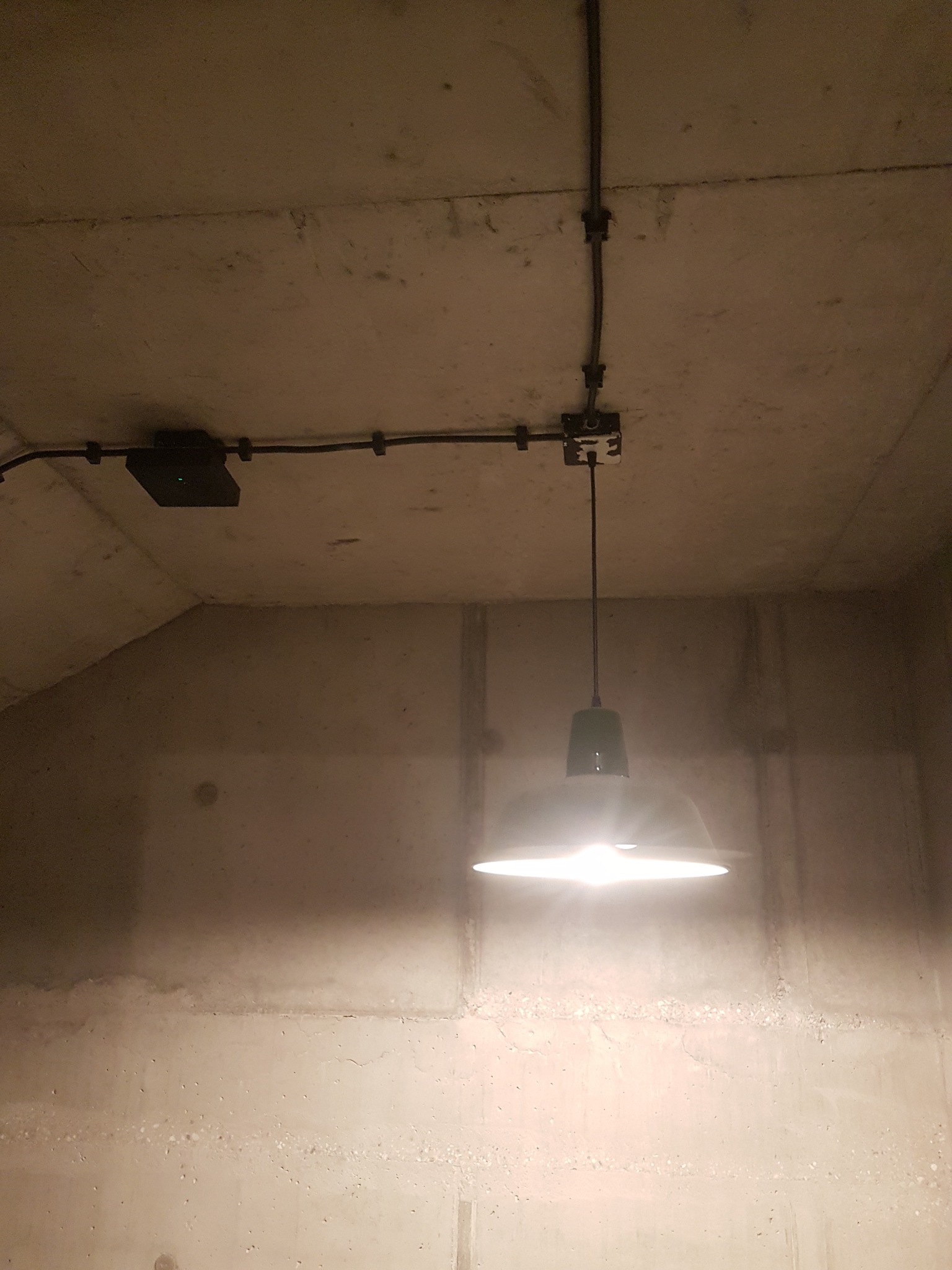 Surowy betonowy sufit z widoczną instalacją elektryczną w czarnych rurkach, puszką połączeniową i zawieszoną lampą z zielonym kloszem, świecącą jasnym światłem.