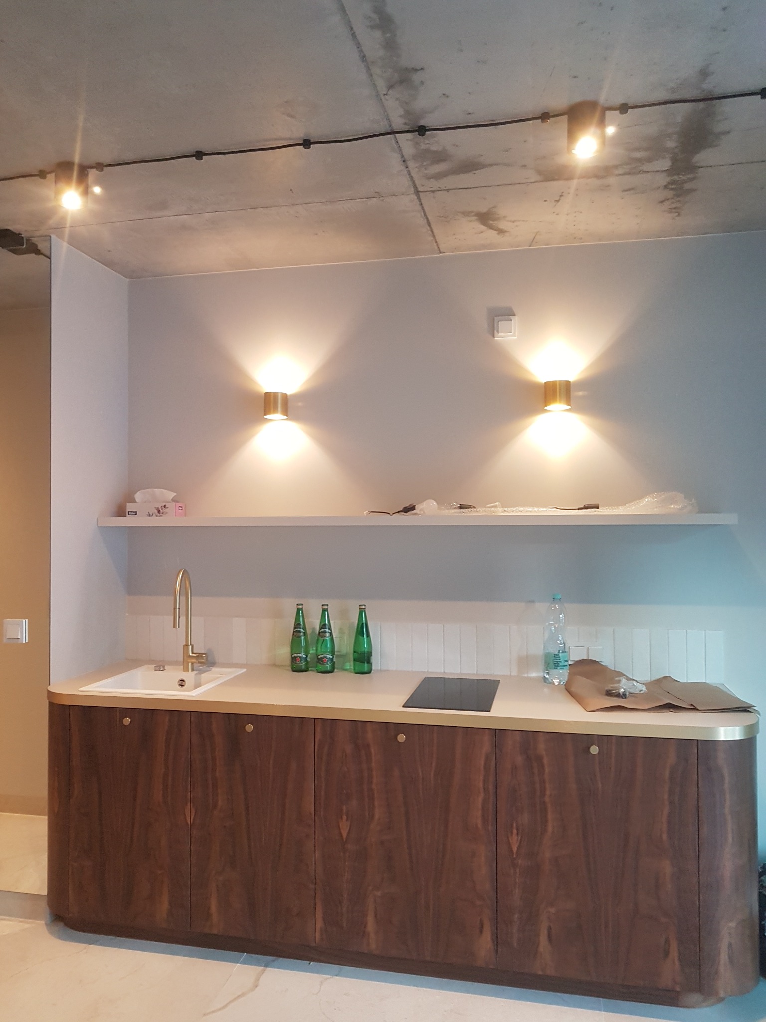 Nowoczesna kuchnia z drewnianymi szafkami, złotą armaturą i oświetleniem punktowym na suficie z betonu architektonicznego, widoczne butelki na blacie i akcesoria montażowe.