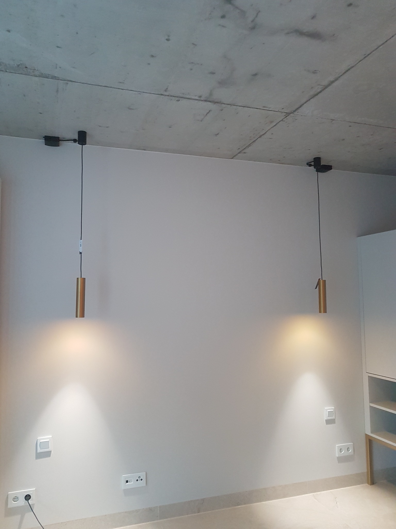 Dwie złote lampy wiszące cylindryczne z czarnymi przewodami zamocowane do surowego betonowego sufitu, widoczne gniazdka elektryczne i włączniki na ścianie.