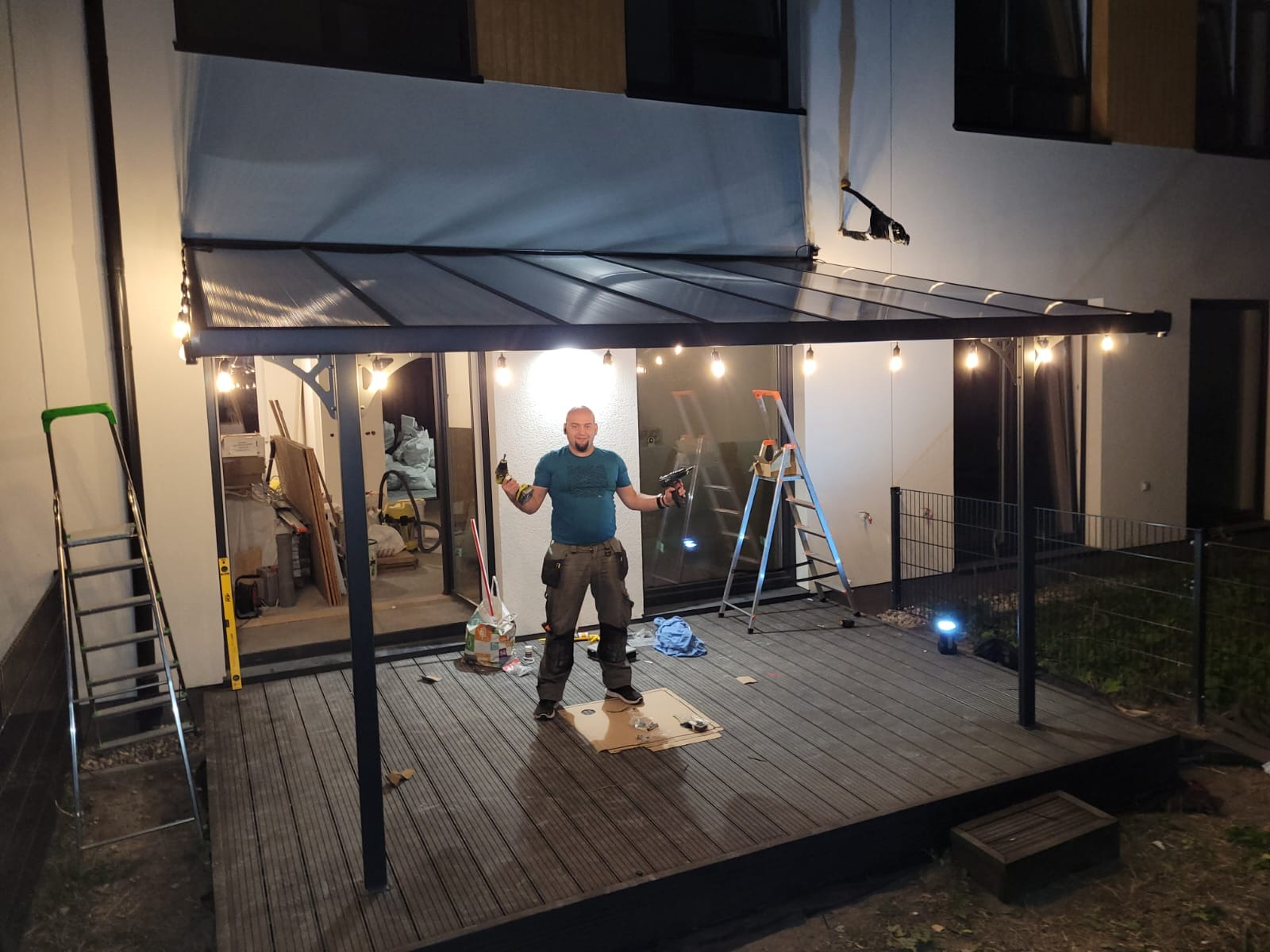 Mężczyzna z wiertarkami w rękach, stojący na kartonie na drewnianym tarasie pod zadaszeniem z oświetleniem, drabiny w tle, prawdopodobnie po montażu konstrukcji.