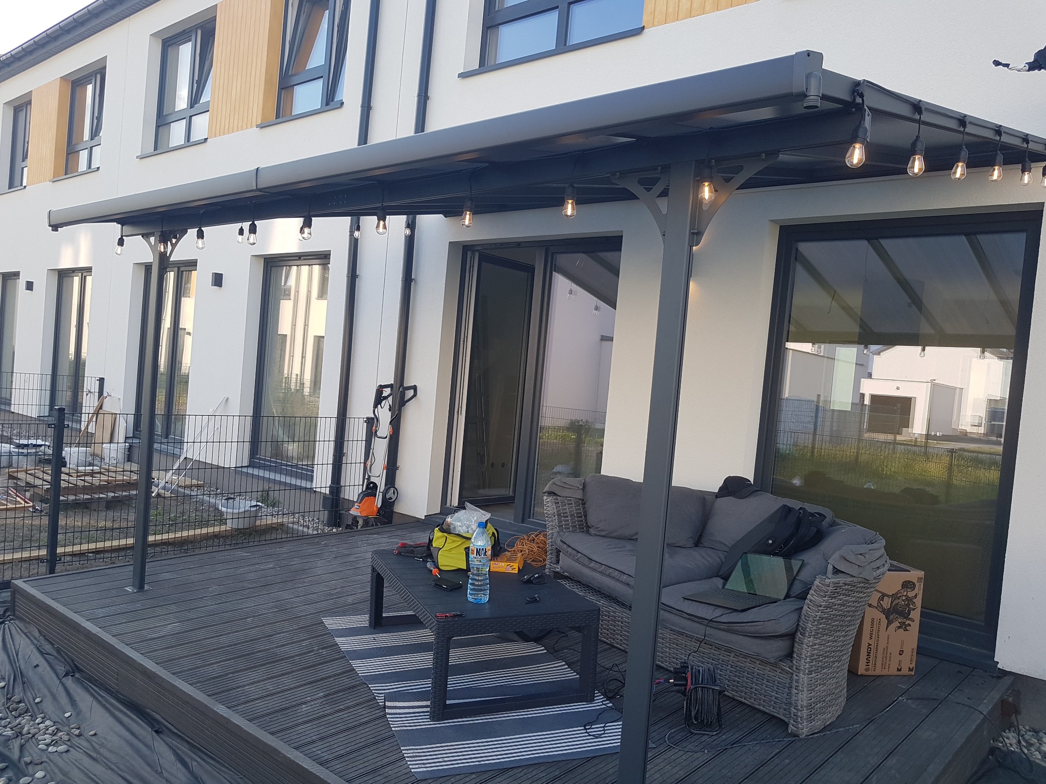 Zadaszony taras z szarą pergolą oświetloną girlandą żarówek, na drewnianej podłodze stolik kawowy z narzędziami, butelką wody i przekąskami, obok sofa z laptopem, w tle nowoczesny dom z białymi...