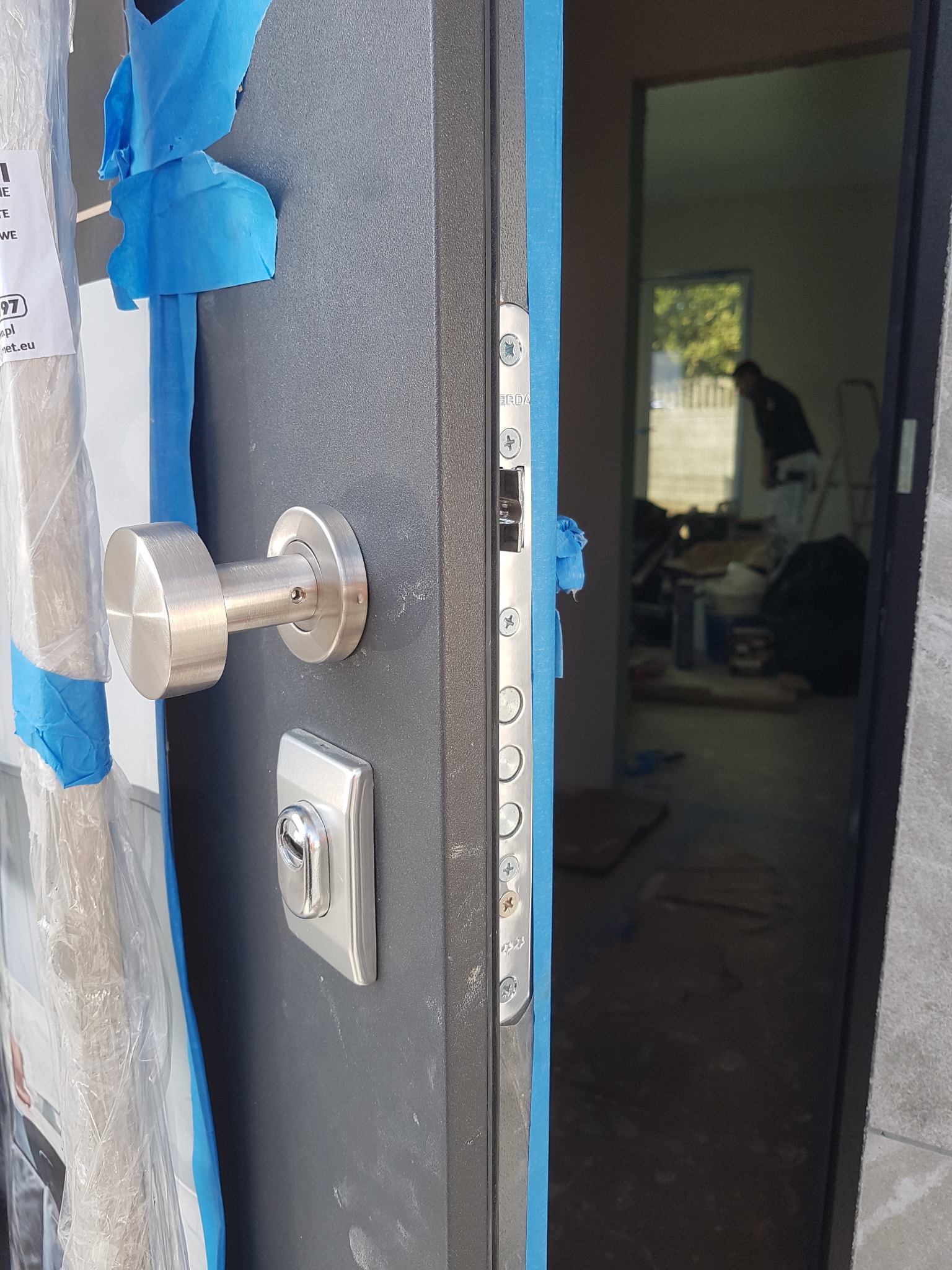 Szare drzwi wejściowe z metalową klamką i zamkiem, zabezpieczone niebieską taśmą malarską, widok na wnętrze w trakcie remontu z sylwetką osoby na tle okna.