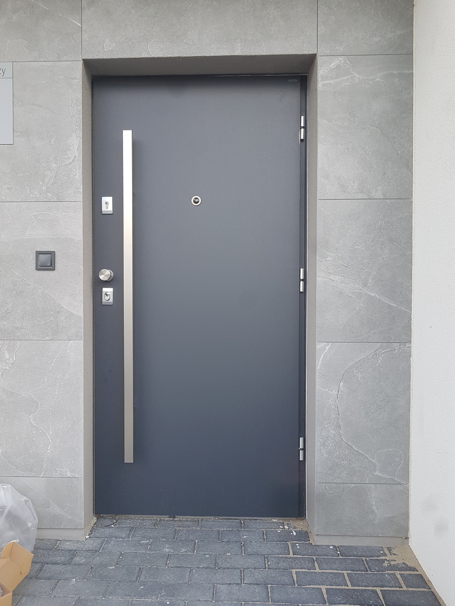Grafitowe drzwi wejściowe z długim, pionowym uchwytem, zamontowane w szarej, kamiennej elewacji, widoczny fragment kostki brukowej i narożnik kartonu.