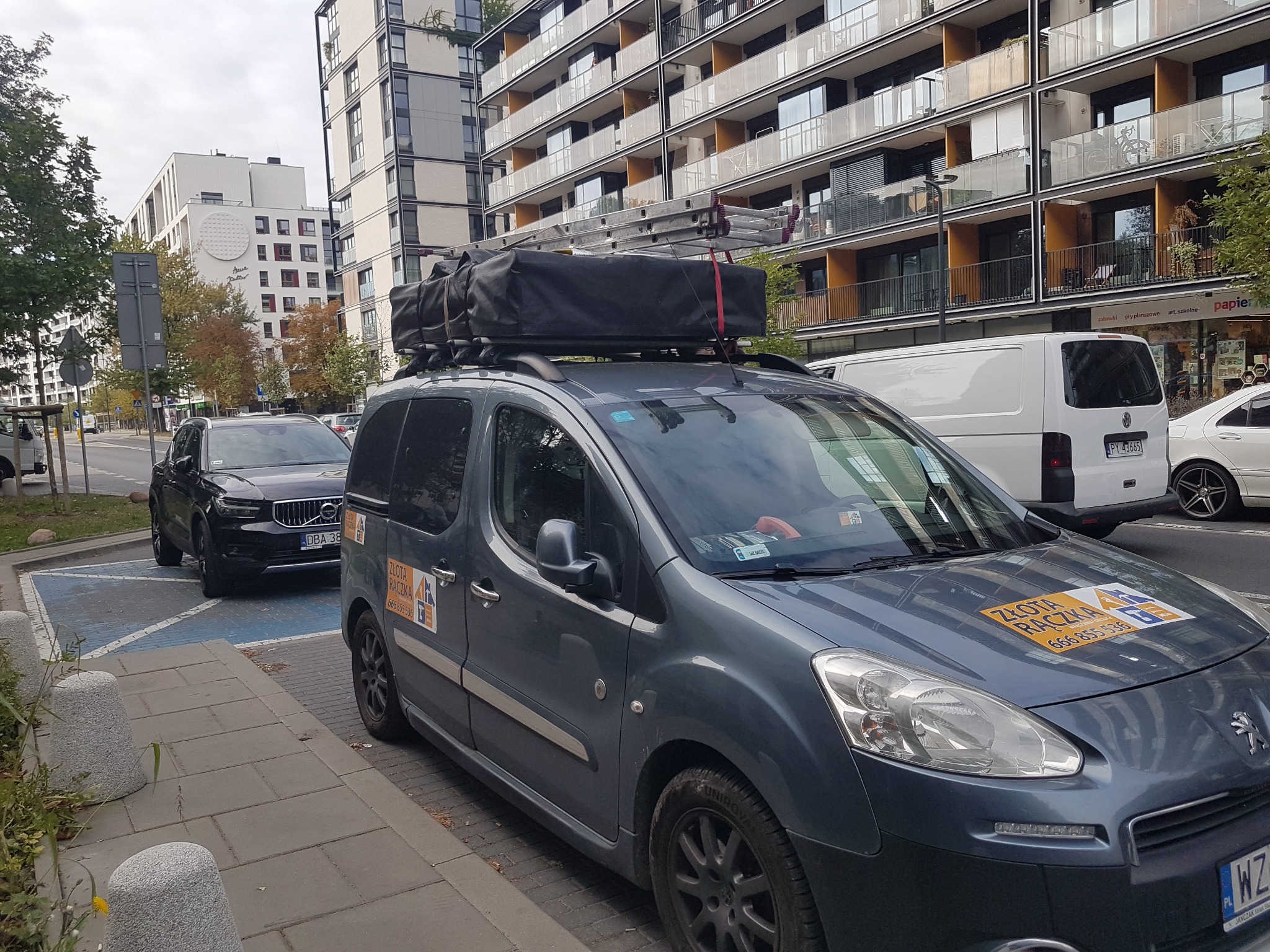 Szary samochód dostawczy z logo firmy usługowej i drabiną na dachu, zaparkowany na ulicy w mieście.