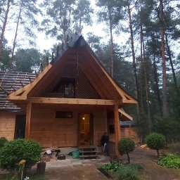 ProjektC24 -Konstrukcje Drewniane Adam Kamiński - Surowy dom szkieletowy w lesie, widoczne elementy konstrukcyjne dachu i drewniane ściany, etap budowy, osoba w drzwiach.