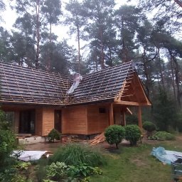 ProjektC24 -Konstrukcje Drewniane Adam Kamiński - Wznoszenie drewnianego domu w lesie, widoczne elementy konstrukcyjne dachu i ściany z desek, obok leżą materiały budowlane.