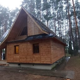 ProjektC24 -Konstrukcje Drewniane Adam Kamiński - Surowy, drewniany dom z wysokim, trójkątnym dachem w lesie, z widocznymi fragmentami dachu pokrytego papą i nieukończonymi elementami wykończeniowymi wokół fundamentów.