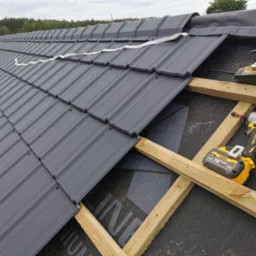 Montaż szarej blachodachówki na drewnianej konstrukcji dachu, widoczne dwa akumulatorowe wkrętaki DeWalt i fragment taśmy uszczelniającej.