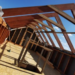 ProjektC24 -Konstrukcje Drewniane Adam Kamiński - Wnętrze w trakcie budowy domu szkieletowego: widok na konstrukcję dachu z belek, płyty OSB na podłodze i czarną membranę na ścianach szczytowych, z fragmentem nieba widocznym przez otwory...