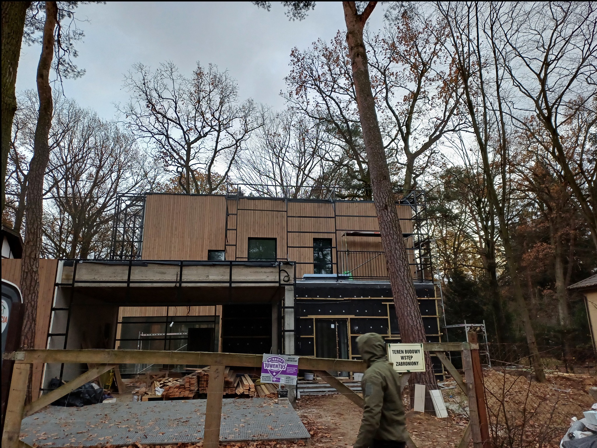 Surowy stan nowoczesnego domu z drewnianą elewacją, widoczne rusztowania i teren budowy w otoczeniu drzew.