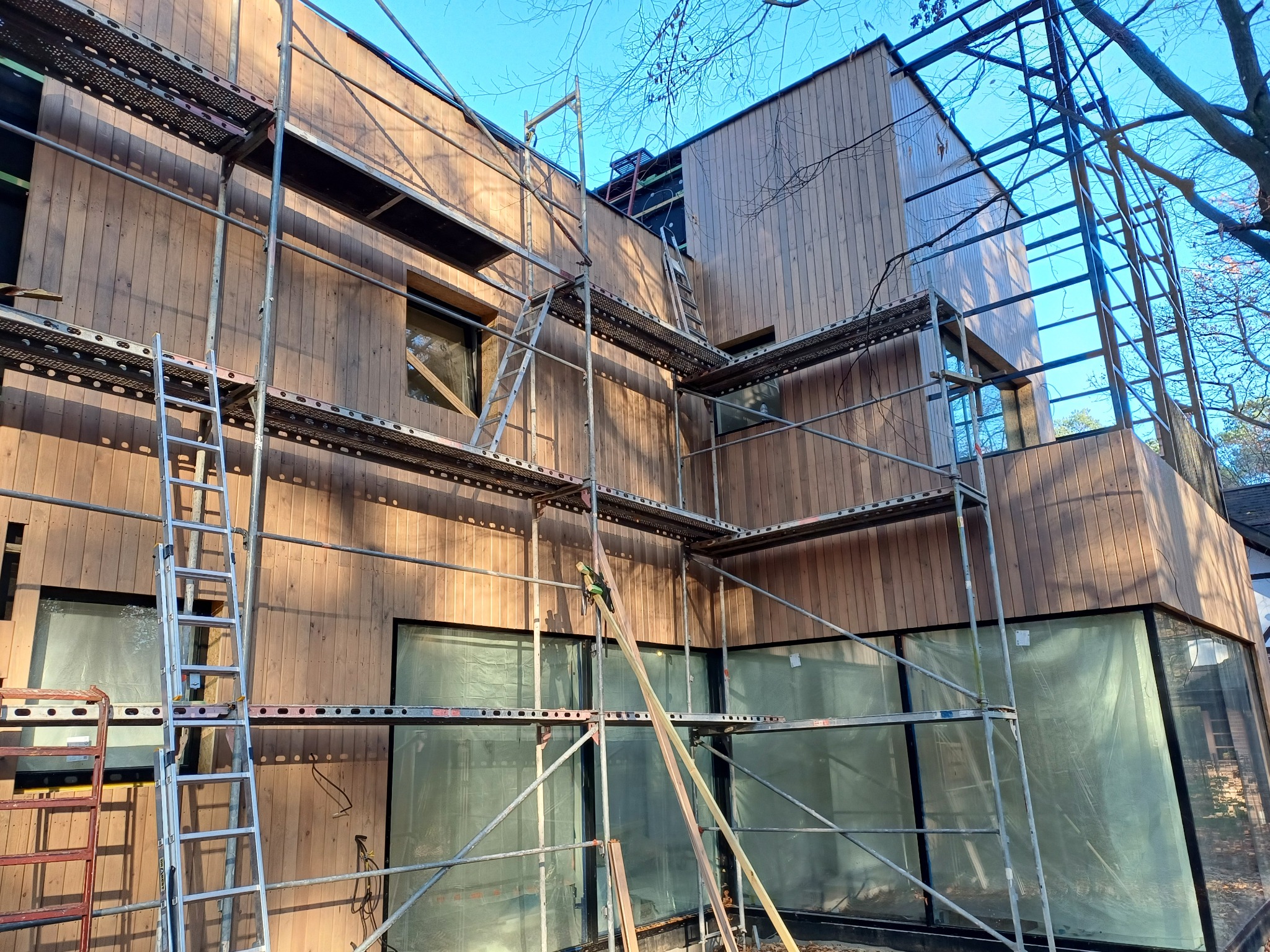 Wznoszenie nowoczesnego budynku z drewnianą elewacją, widoczne rusztowania i zabezpieczone folią okna.