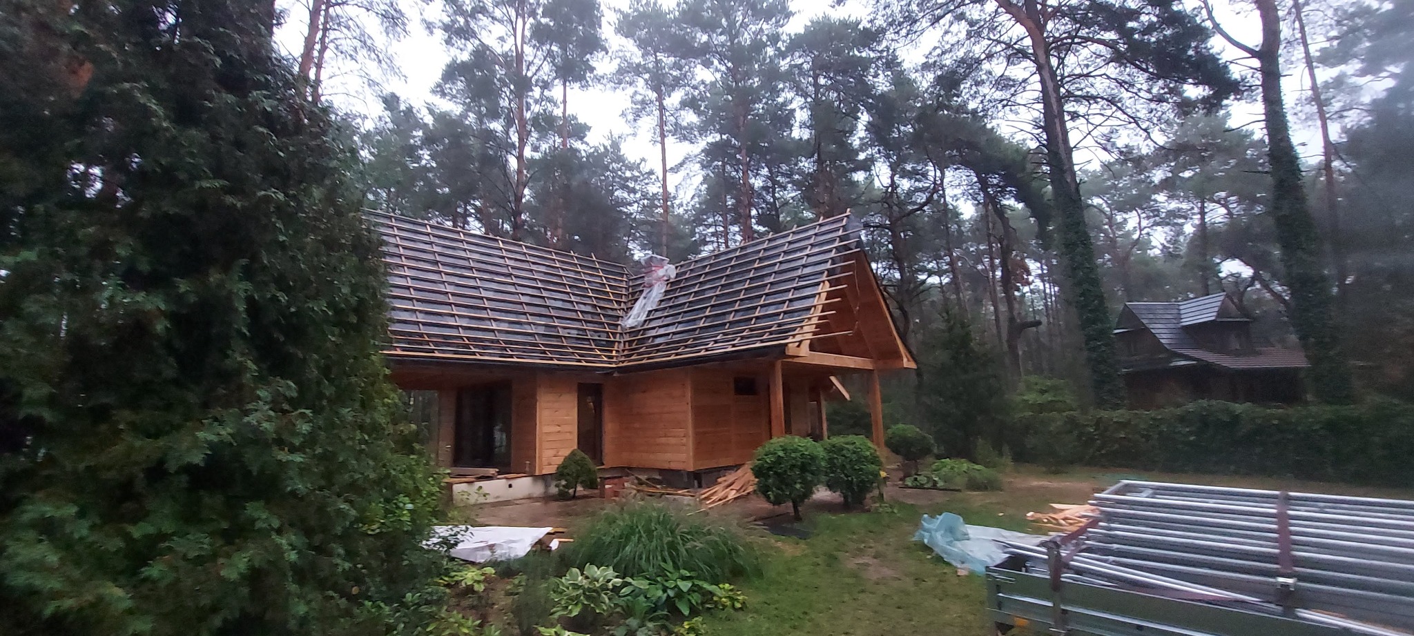 Wznoszenie drewnianego domu w lesie, widoczne elementy konstrukcyjne dachu i ściany z desek, obok leżą materiały budowlane.